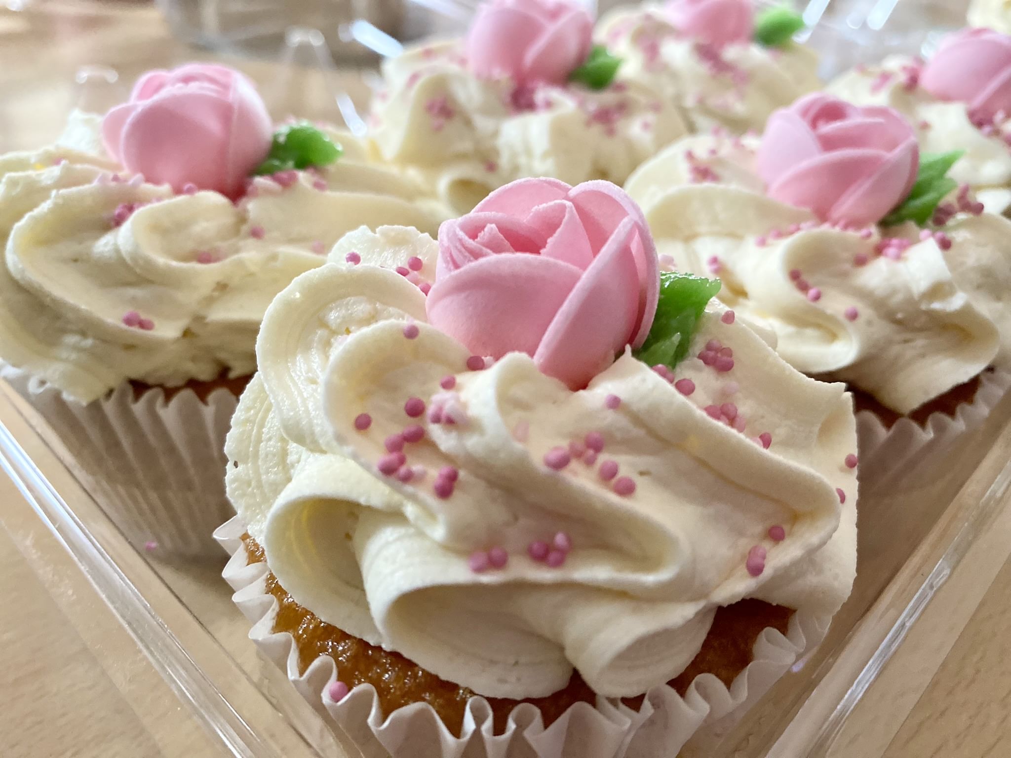 Close-up of cupcakes topped with creamy white frosting, pink rose-shaped decorations, and pink sprinkles. The cupcakes are arranged together in a clear plastic tray.